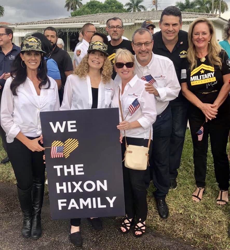 Volunteers in Parkland Cares shirts hold a sign reading “We love the Hixon family” at a Military Makeover event, carrying small American flags.